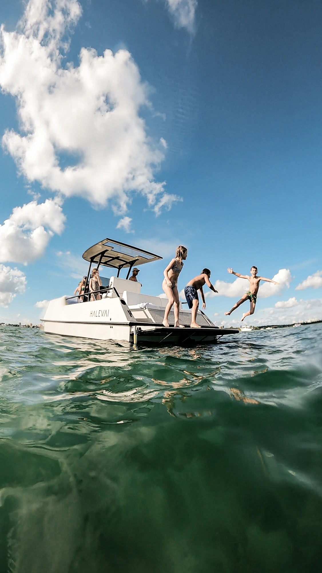 Children jumping off the back platform of a white boat named HALEVA into green water under a blue sky with scattered clouds.
