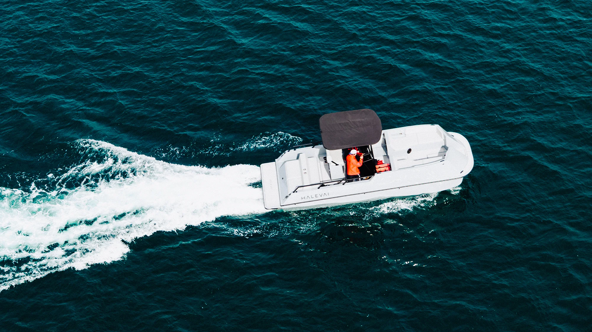 White motorboat with a black canopy and a person in an orange jacket steering on dark blue water, creating a white wake.