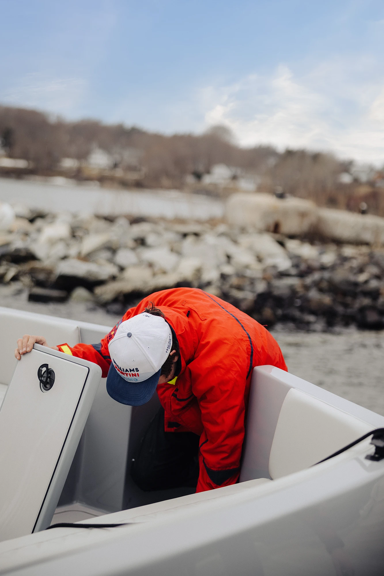 Person wearing a white and blue cap and bright orange jacket bending down inside a white boat near a rocky shoreline.