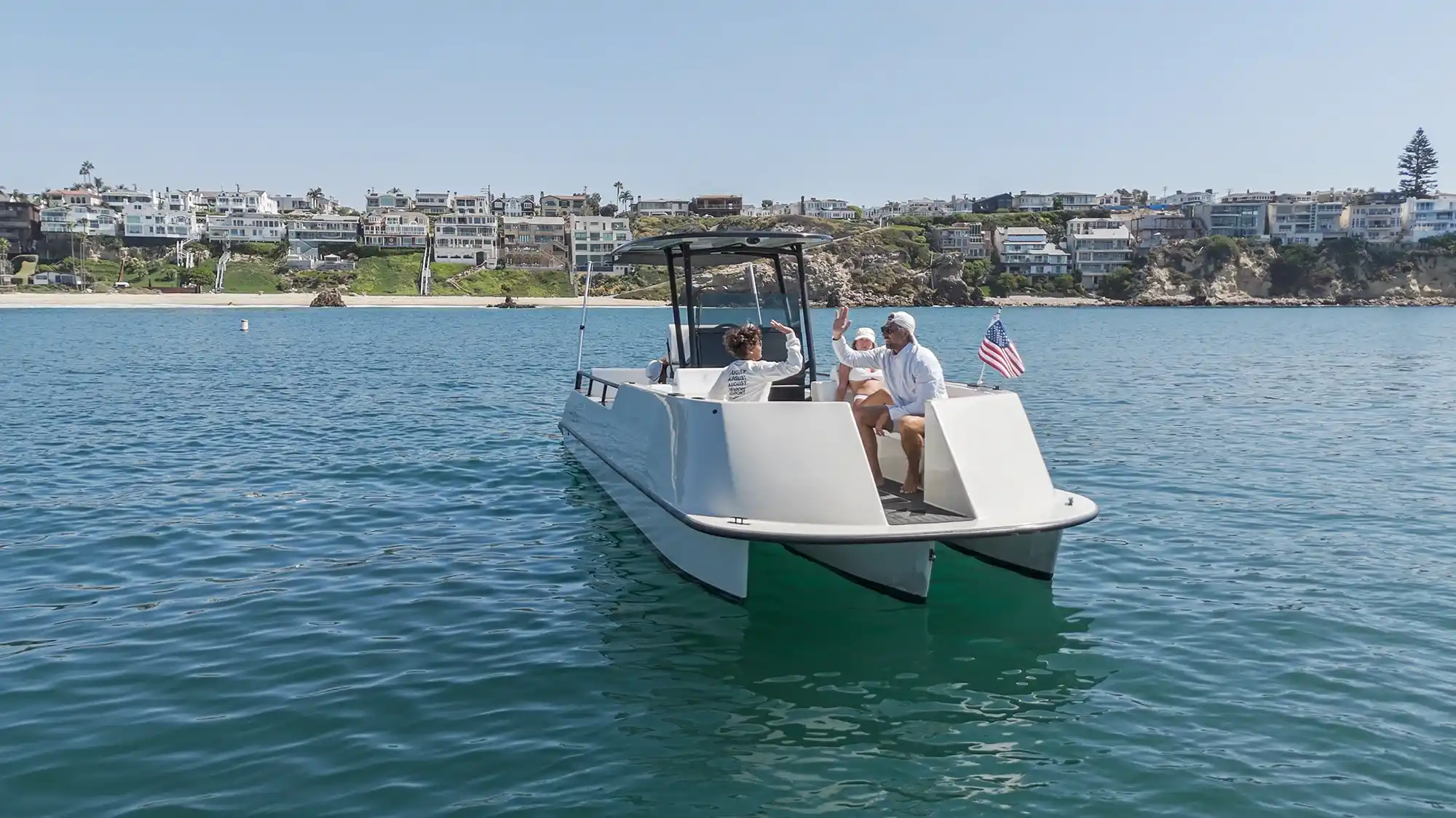 A modern white catamaran boat floating on calm blue water near a shoreline with houses and greenery, with three people on board, two giving a high five and one seated between them, and an American flag at the back.