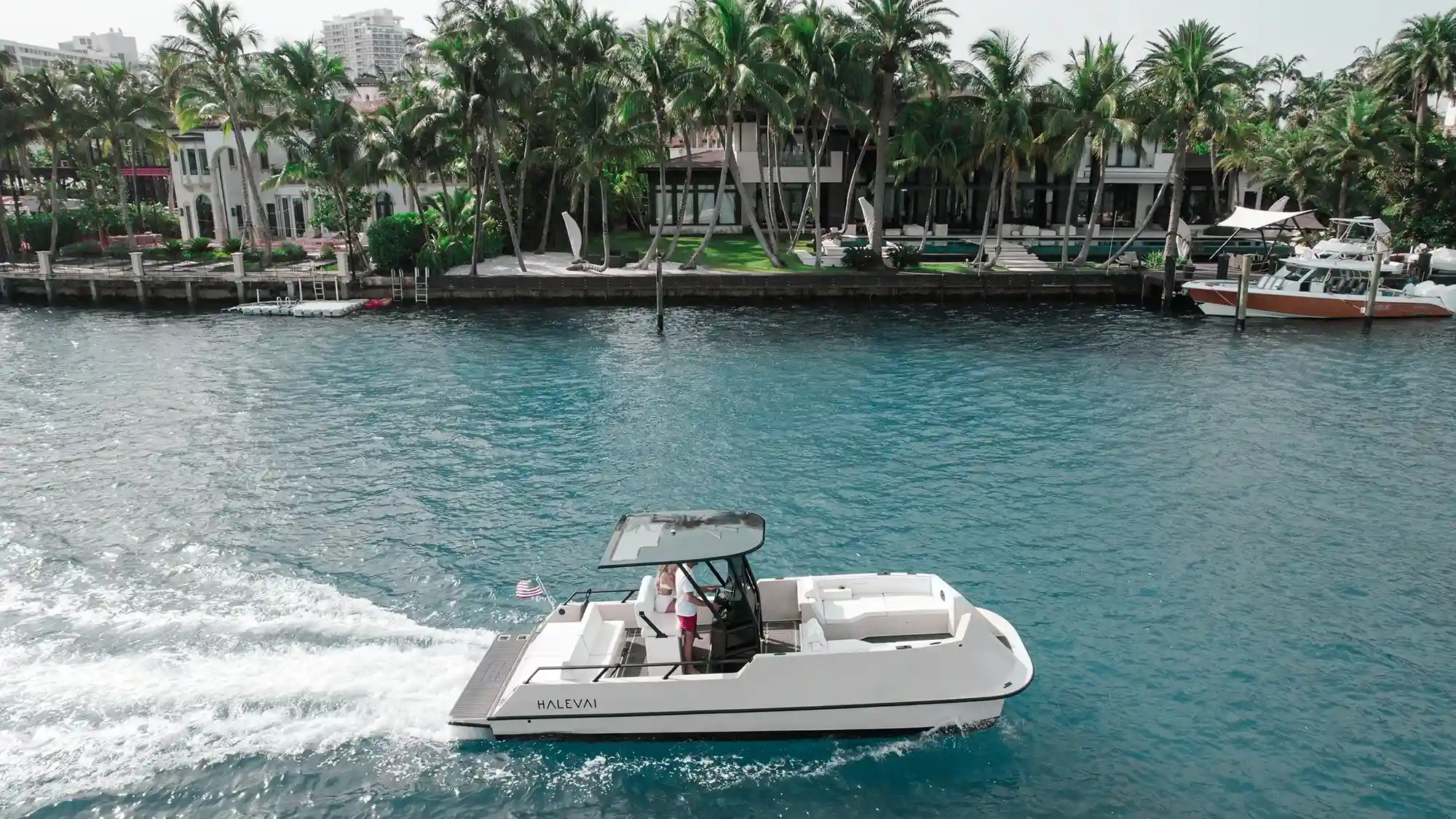 White motorboat named HALEVA I with a canopy cruises on clear blue water near a shore lined with palm trees and modern waterfront houses.