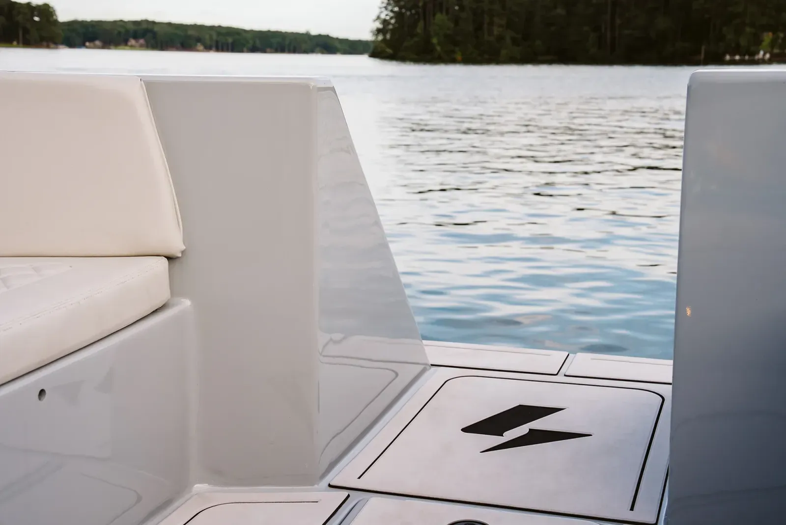 Close-up view of a boat deck with white cushioned seating and a hatch featuring a black lightning bolt symbol, on a calm lake with trees in the background.