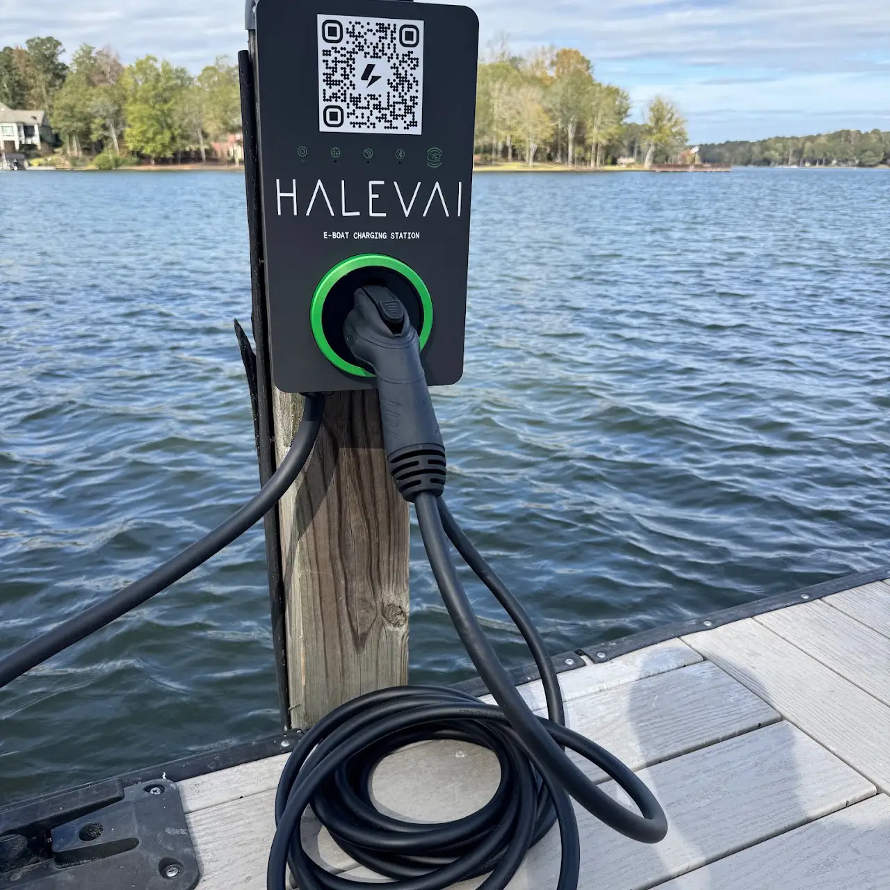 E-boat charging station mounted on a wooden post at a dock by a lake, with a black charging cable coiled on the dock and extending into the station.