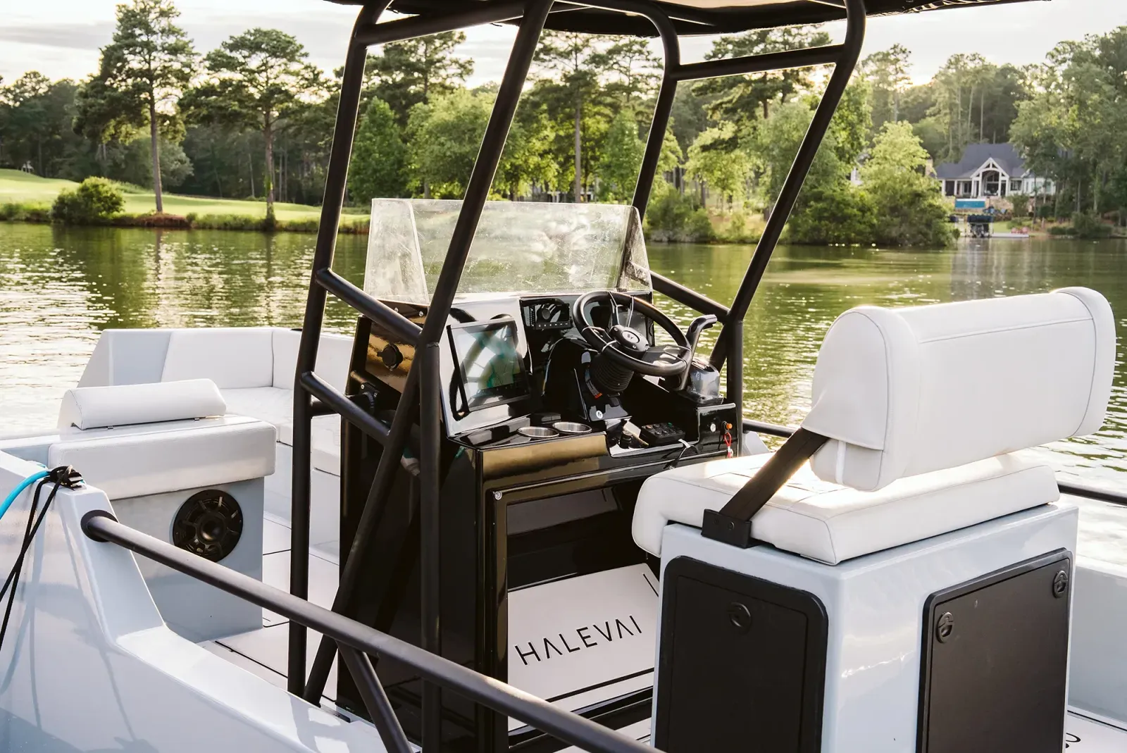 The helm of a small modern boat with a white cushioned captain's seat, black steering wheel, control panel, and touchscreen display, set against a lake with green trees and houses in the background.