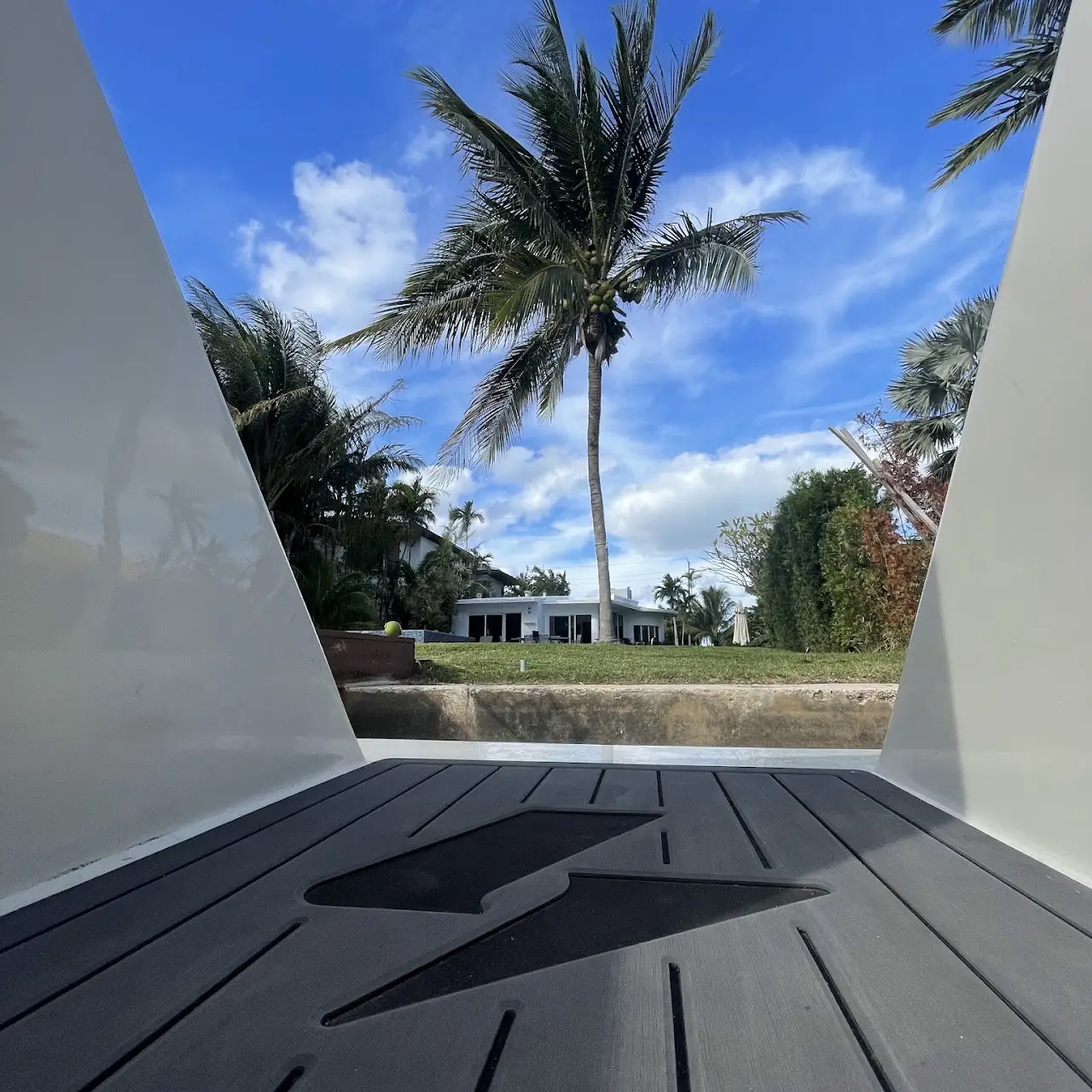 View over a boat's deck with a lightning bolt symbol looking towards a yard with a tall palm tree, bushes, and a white house under a blue sky with scattered clouds.