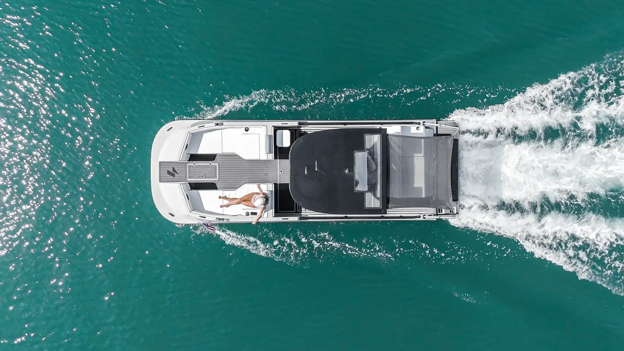 Aerial view of a white motorboat cruising on teal water with a person in a white swimsuit lying on its deck near the rear.