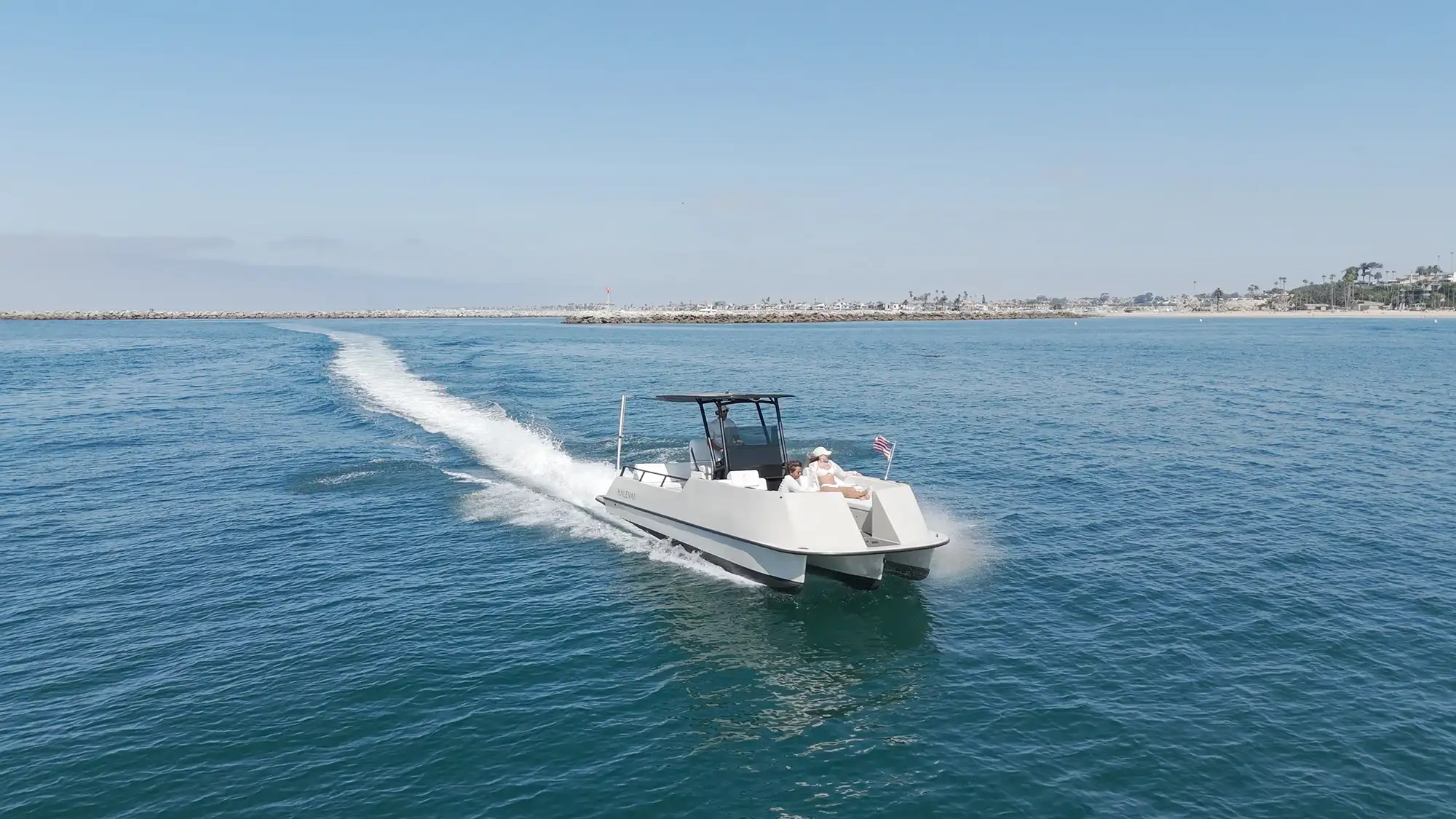 White motorboat with two people cruising on calm blue water near a coastal area with a sandy beach and scattered buildings under a clear blue sky.