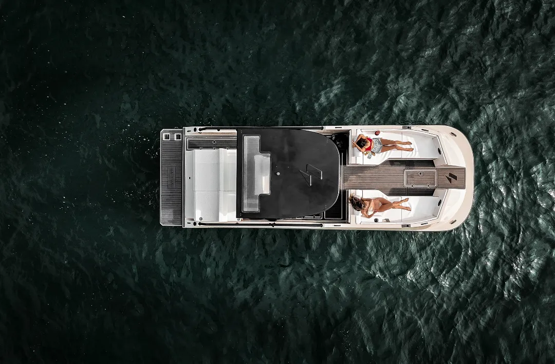Aerial view of a small modern boat floating on dark water with two people lying on the bow sunbathing, one wearing red and the other white.