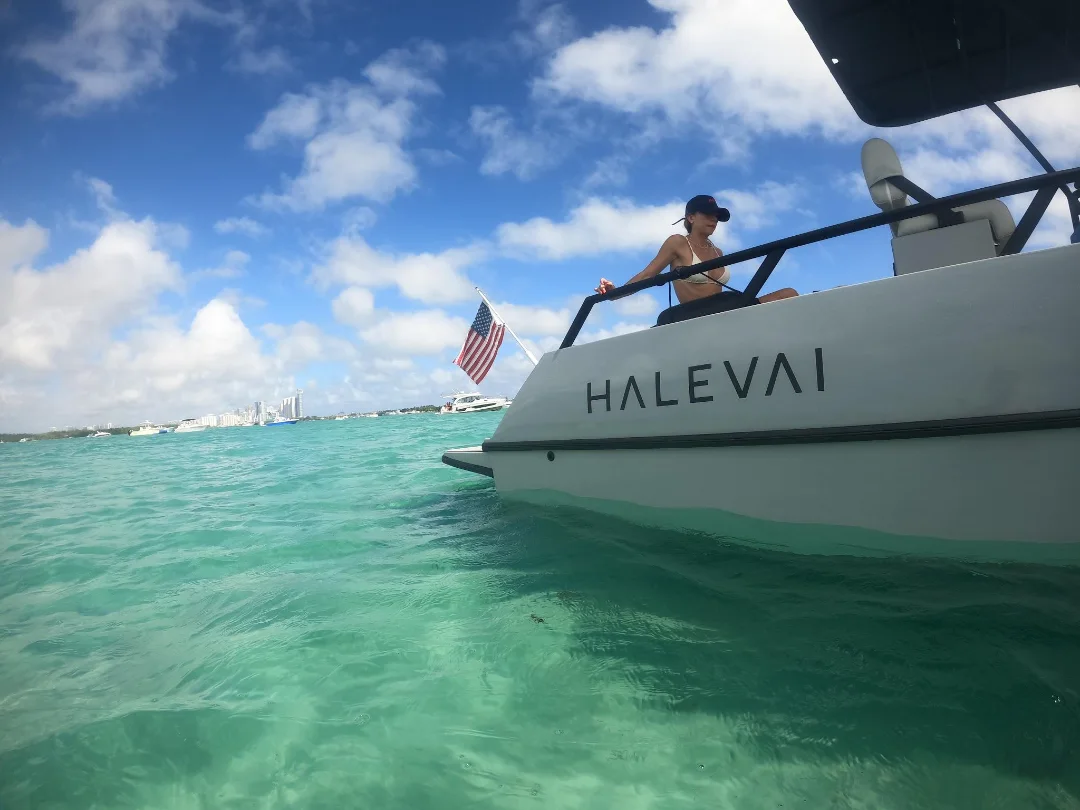 Woman in a bikini and cap sitting on the edge of a white boat named HALEVAI floating on clear turquoise water with an American flag at the stern under a partly cloudy blue sky.