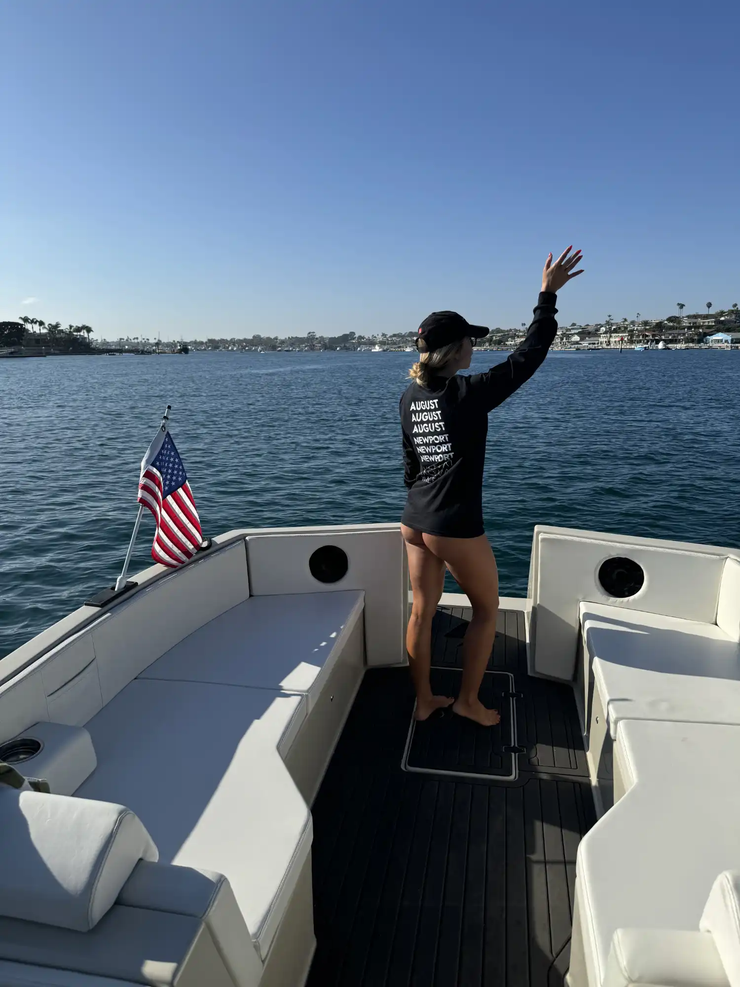 Person wearing a black long-sleeve shirt and cap, standing barefoot on the deck of a boat and waving towards a waterfront with houses and palm trees under a clear blue sky.