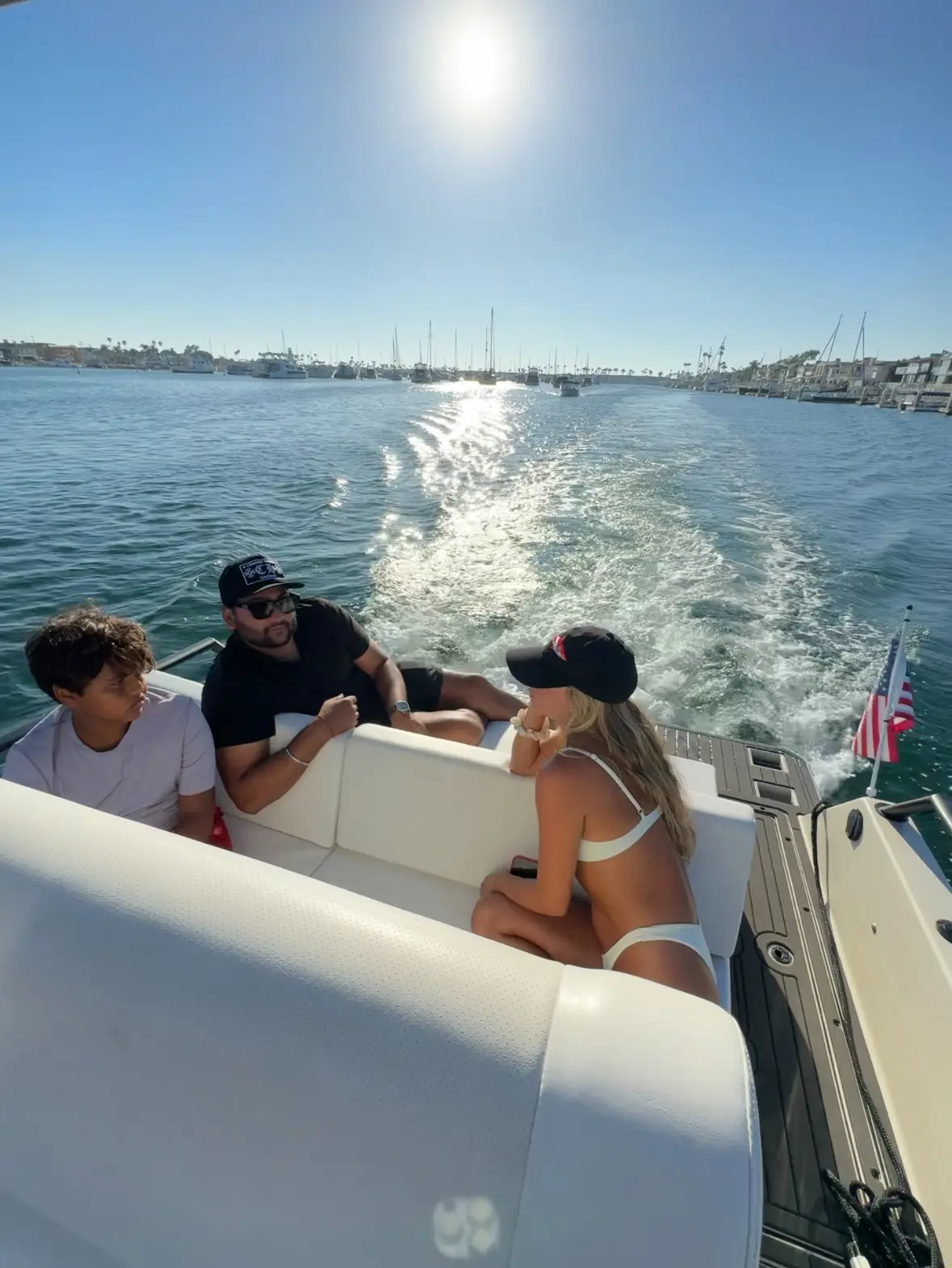 Three people relaxing on a white boat with an American flag at the stern, cruising on a calm waterfront marina under a bright sun and clear blue sky, with many docked sailboats in the distance.