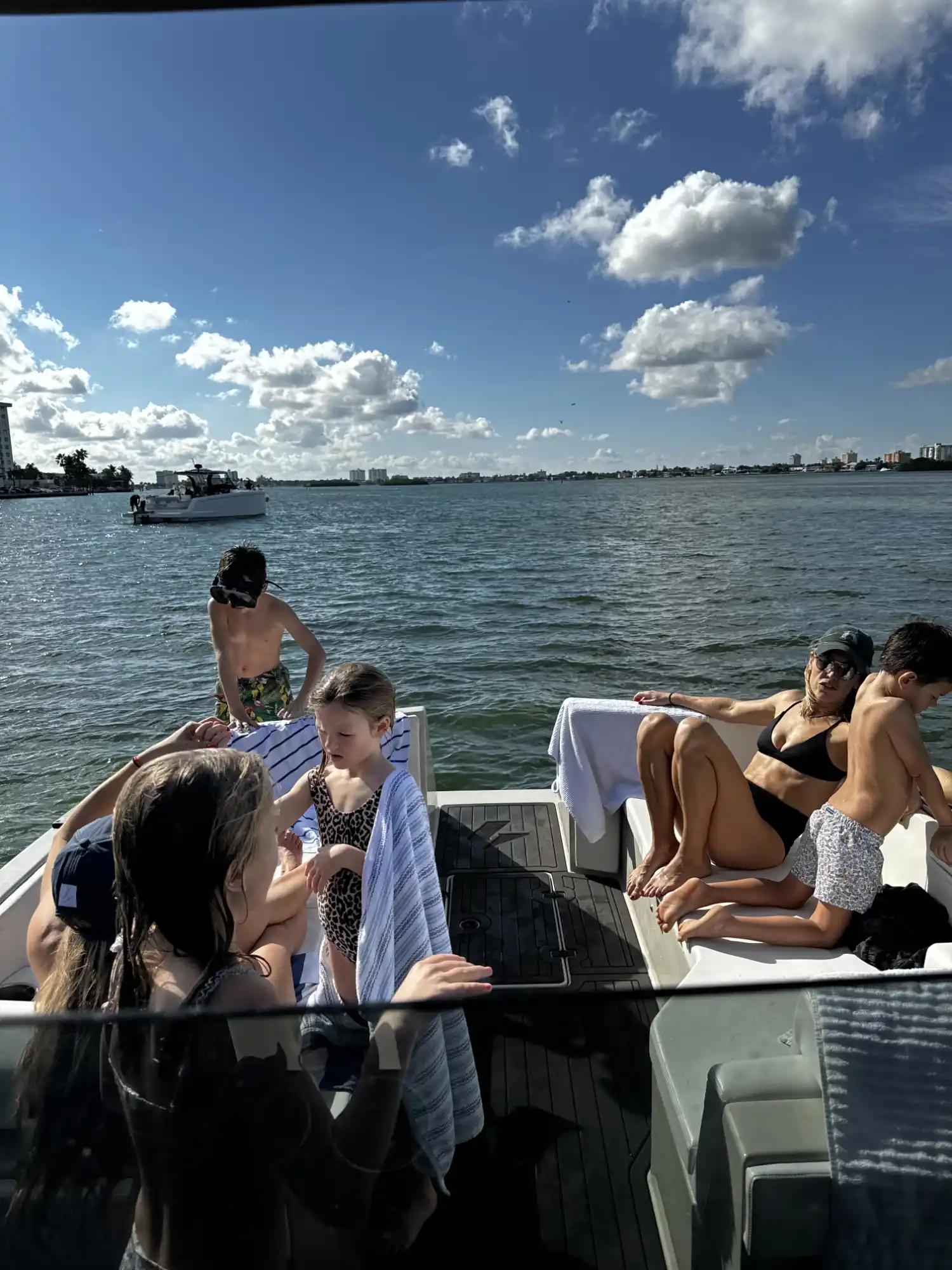 Group of people, including children and an adult woman in a black bikini and sunglasses, relaxing and interacting on a boat with water and a partly cloudy sky in the background.