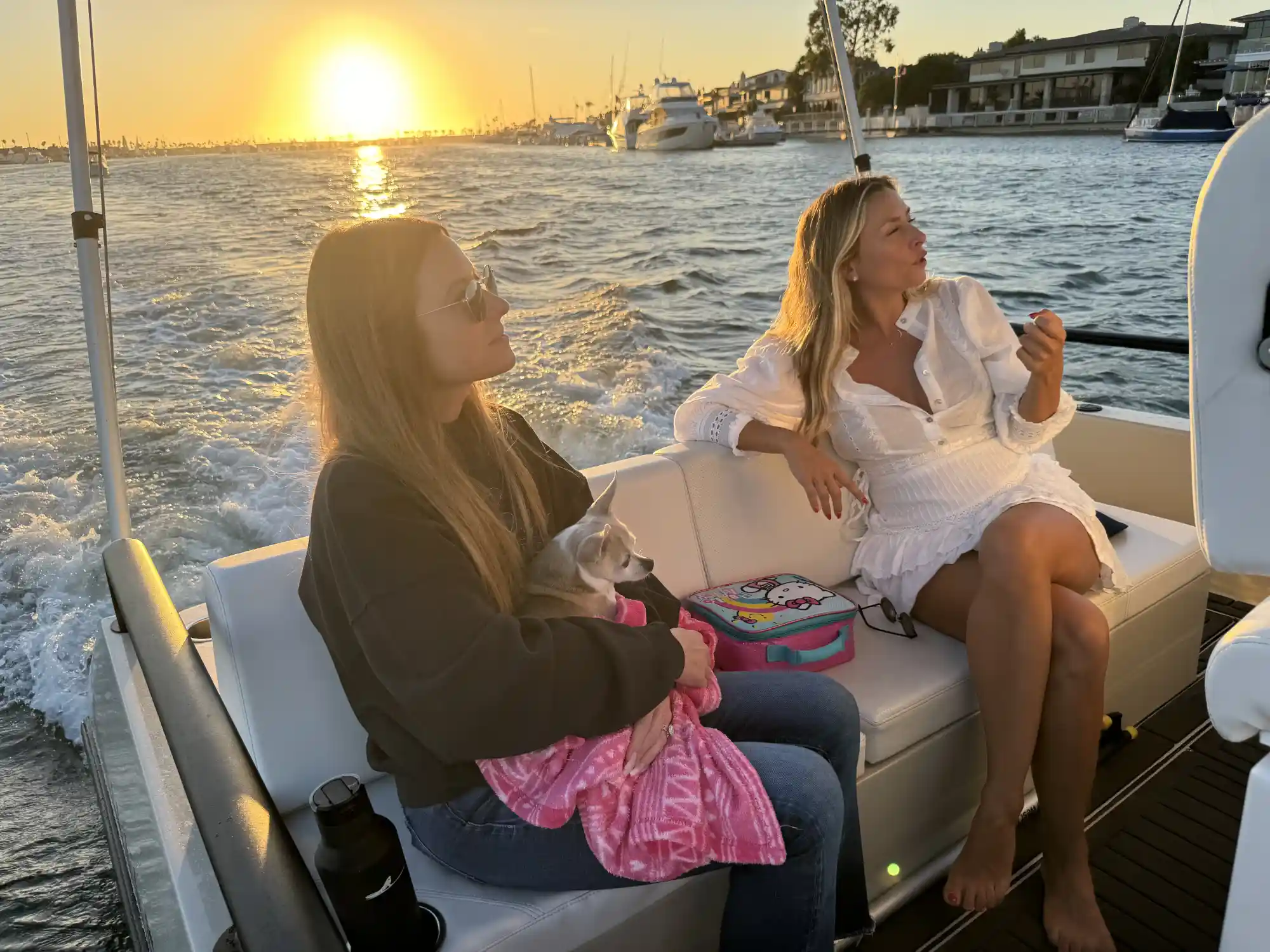 Two women sitting on a boat during sunset, one holding a small dog wrapped in a pink blanket, with water and boats in the background.