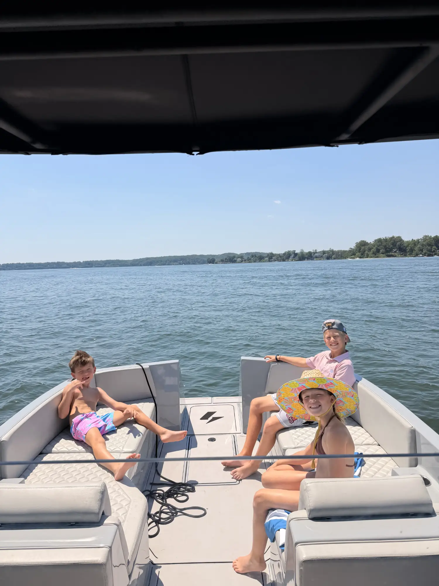 Three children sitting and relaxing on the cushioned seats of a boat on a sunny day, with calm water and a tree-lined shore in the background.