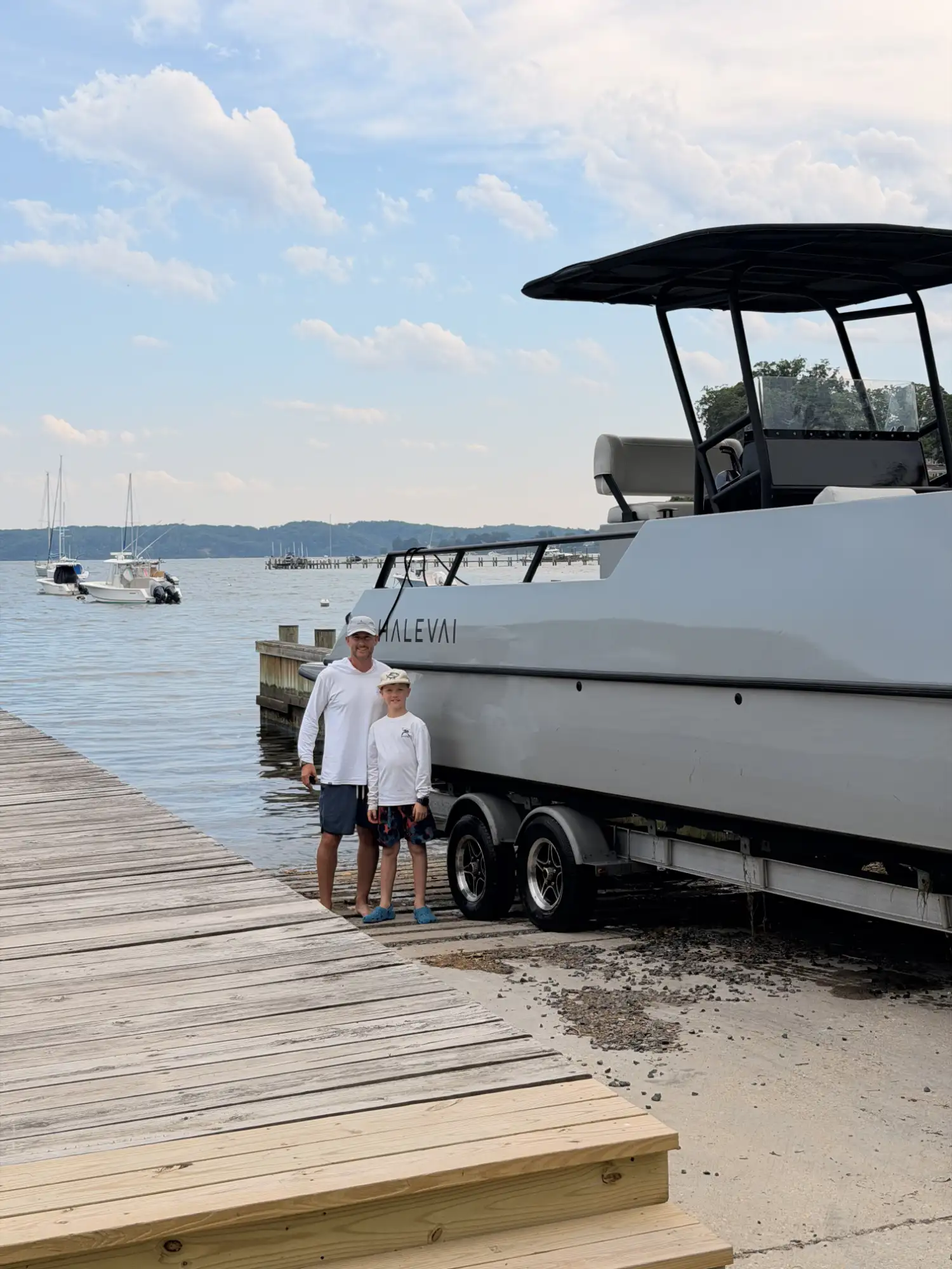 Man and boy wearing white long-sleeve shirts and hats standing next to a large gray boat on a trailer by a wooden dock with water and sailboats in the background under a partly cloudy sky.