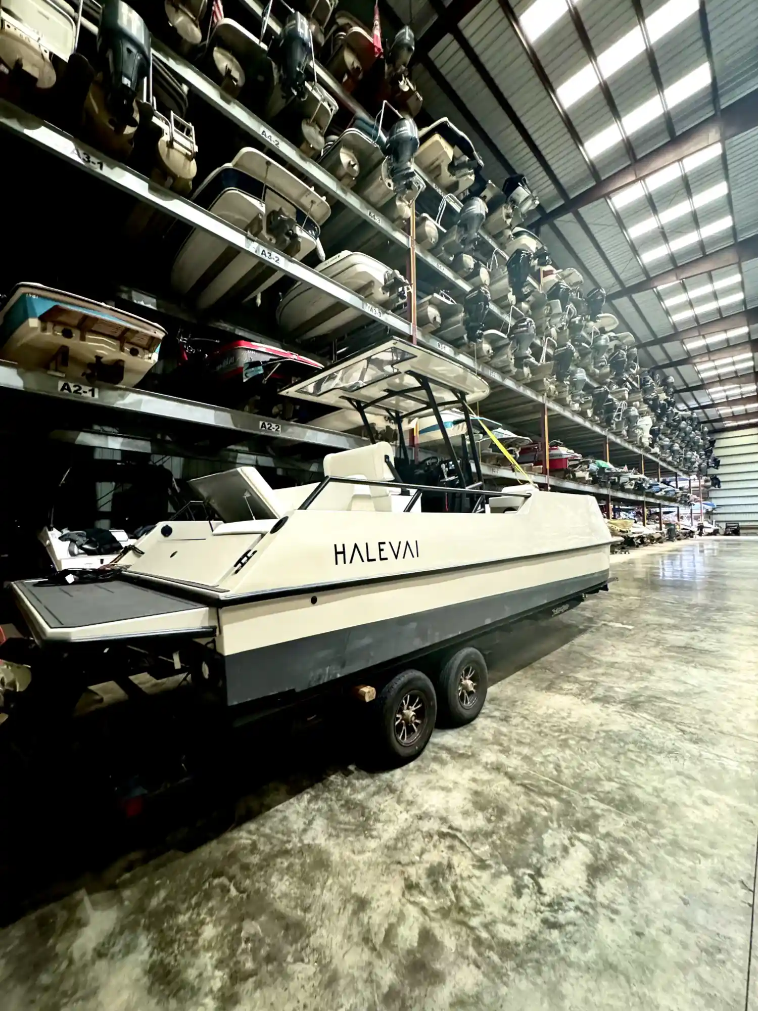 White motorboat named HALEVAI on trailer inside a large warehouse with multiple racks holding many boats stacked in rows.