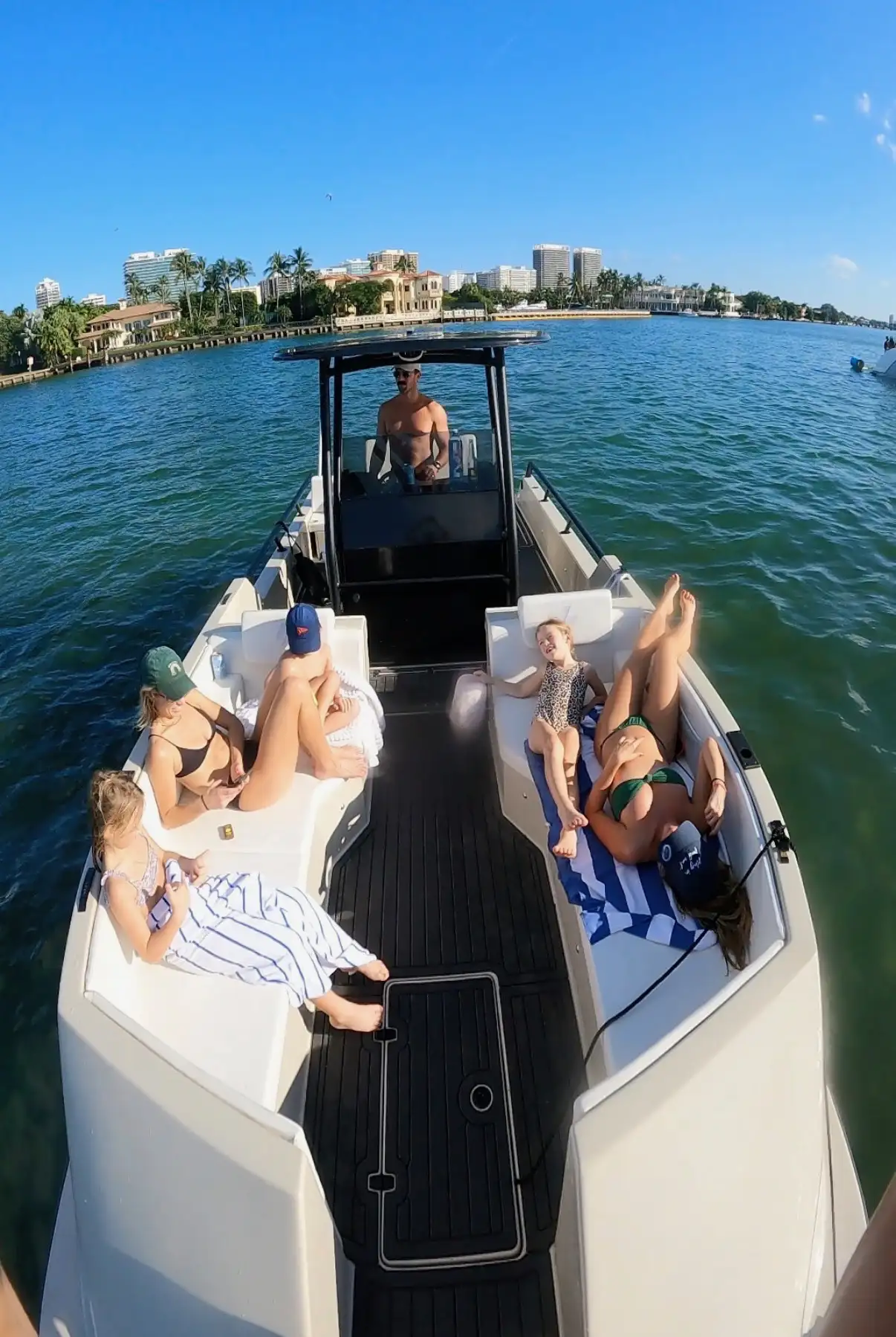 A man steering a boat on water with a shirtless upper body, while five females of varying ages relax on the boat's seats under a clear blue sky with a city skyline and palm trees in the background.