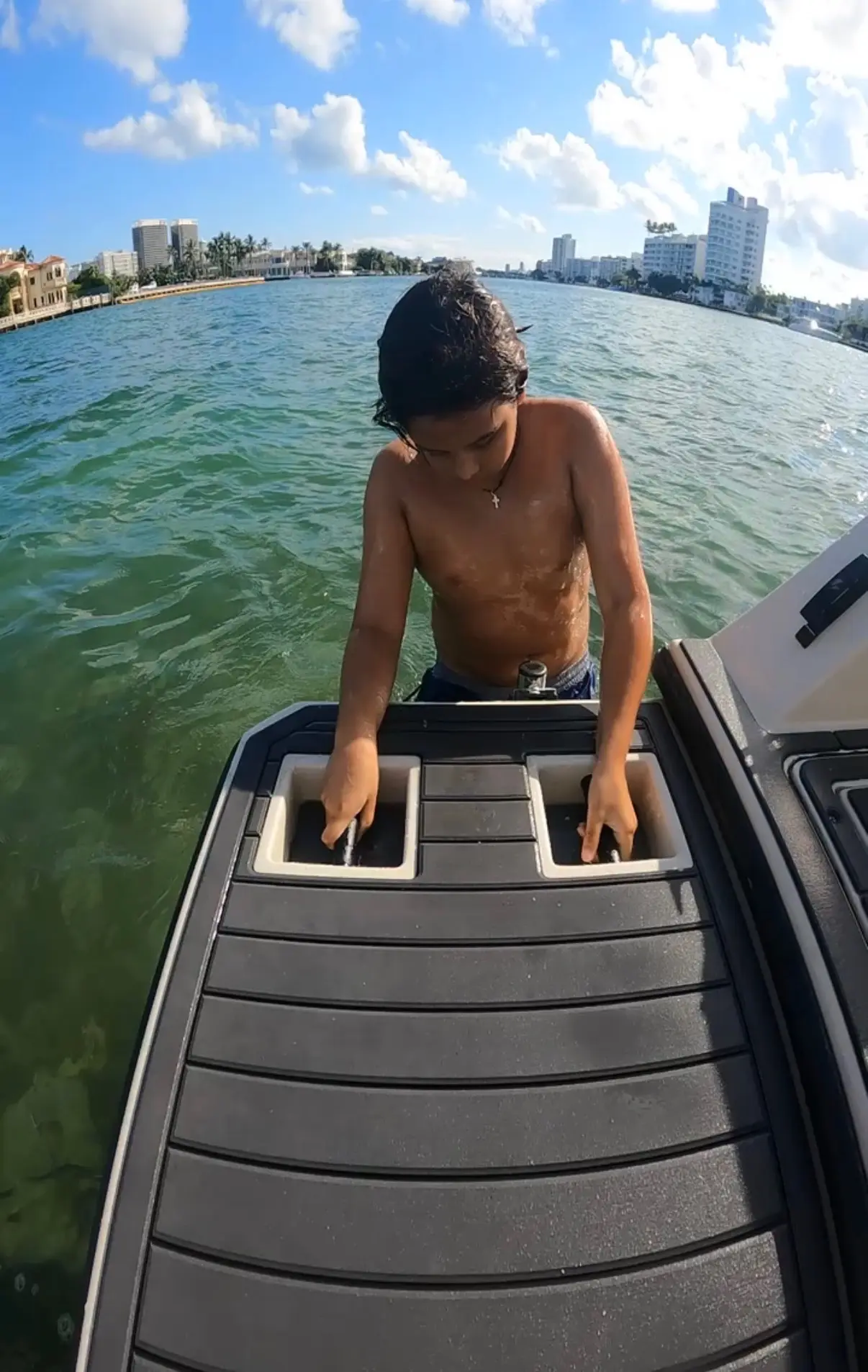 A shirtless boy with wet hair and a cross necklace is on a boat, holding two handles set in a black surface with water and city buildings in the background under a partly cloudy sky.