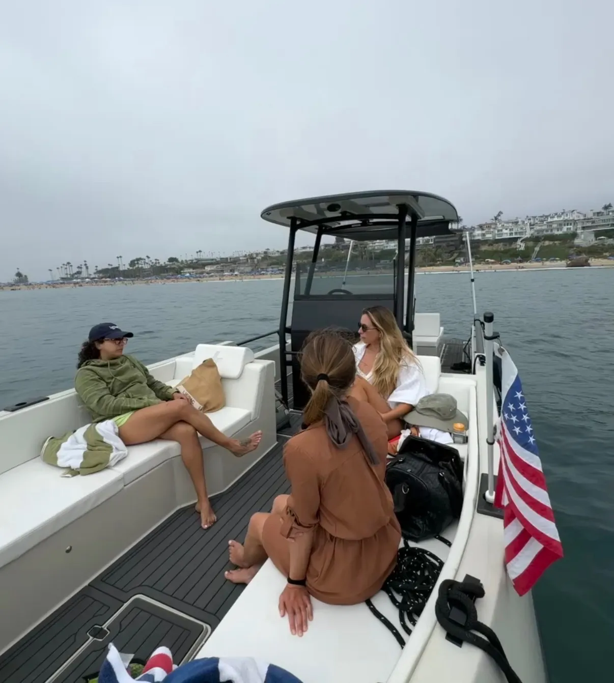 Three women relaxing on a boat near the coast, with one wearing a green hoodie and cap sitting on the left, another with long blonde hair in white sitting towards the front, and the third with a brown dress and hair tied back sitting in the center facing forward. An American flag is attached to the right side of the boat, and the shore with buildings and palm trees is visible in the background under a cloudy sky.