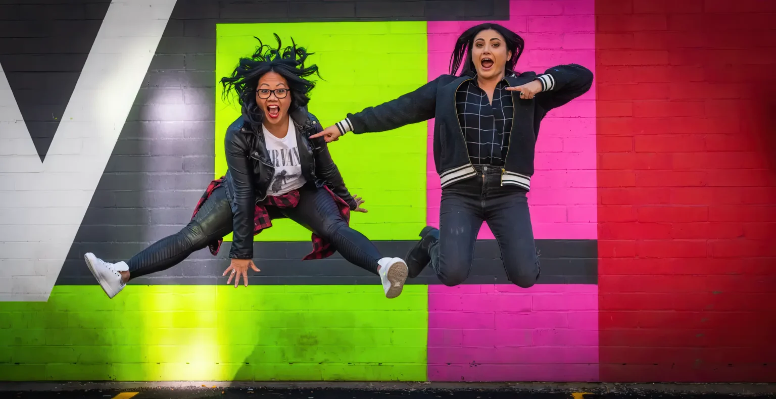 Two community members jumping together in front of a colorful wall.