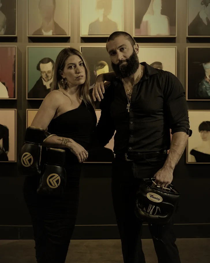 A woman in a black one-shoulder dress holds boxing gloves beside a man in a black shirt holding headgear, in front of a wall of portraits