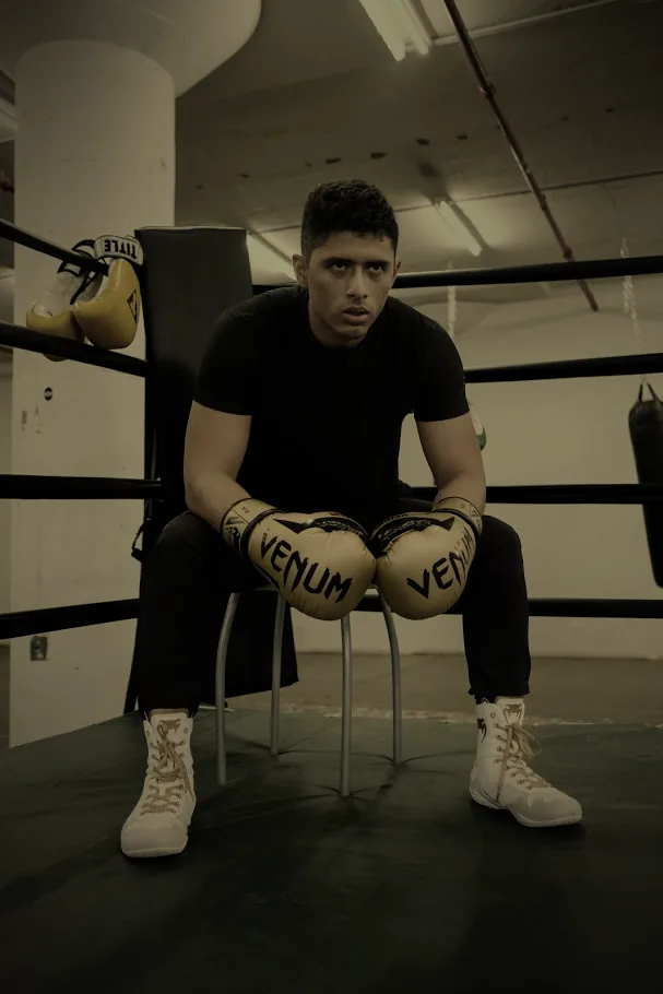 A boxer in gold gloves sits on a stool in the corner of the ring