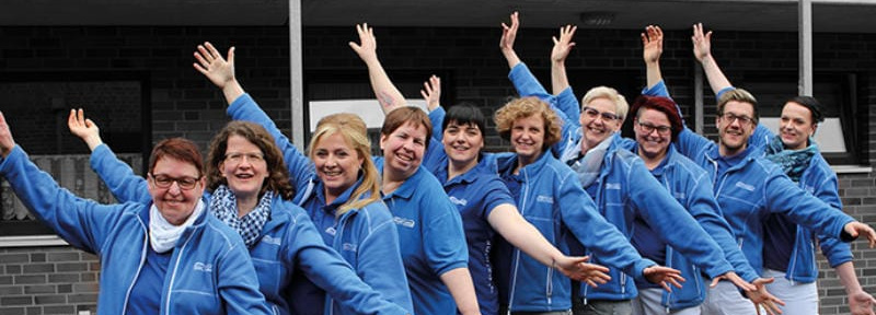 A cheerful Buurtzorg team in blue jackets waving with their arms raised