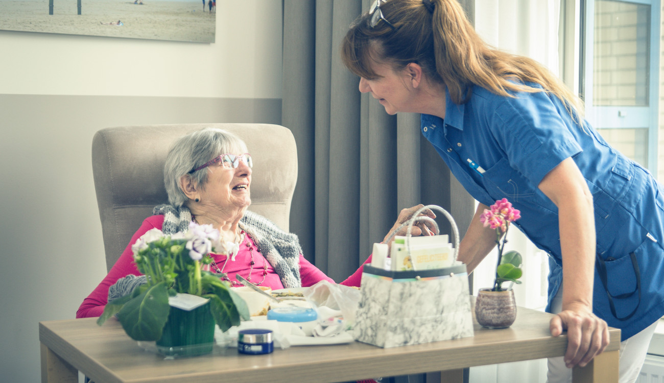 A carer leaning in to talk with an older woman seated at a table with flowers