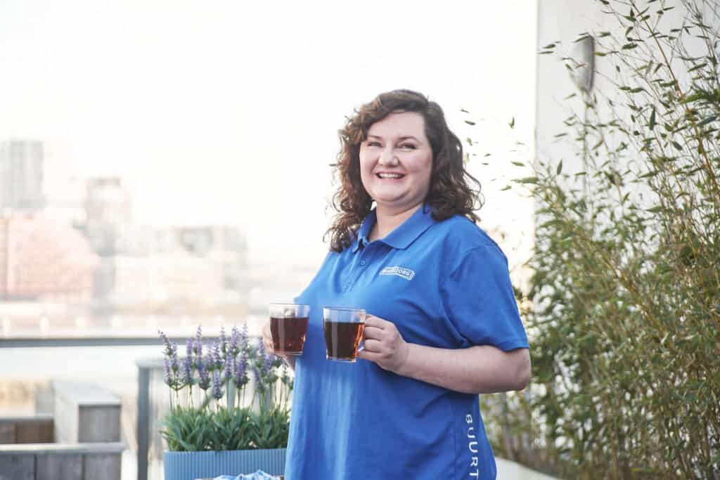 A smiling woman in a blue Buurtzorg polo shirt carrying two glasses of tea