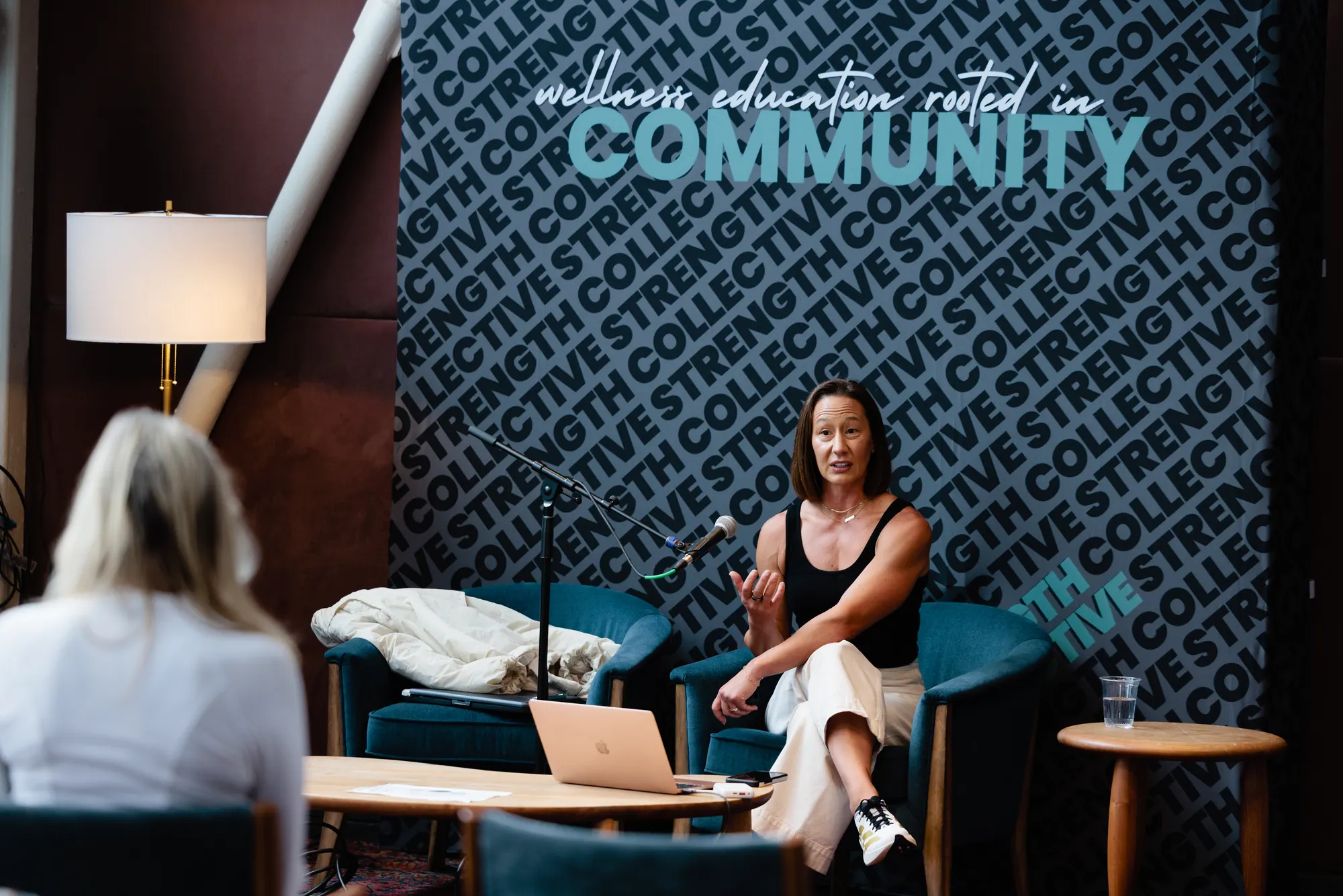 A speaker in a teal armchair talking into a microphone, a laptop on the table beside her and the audience in the foreground.