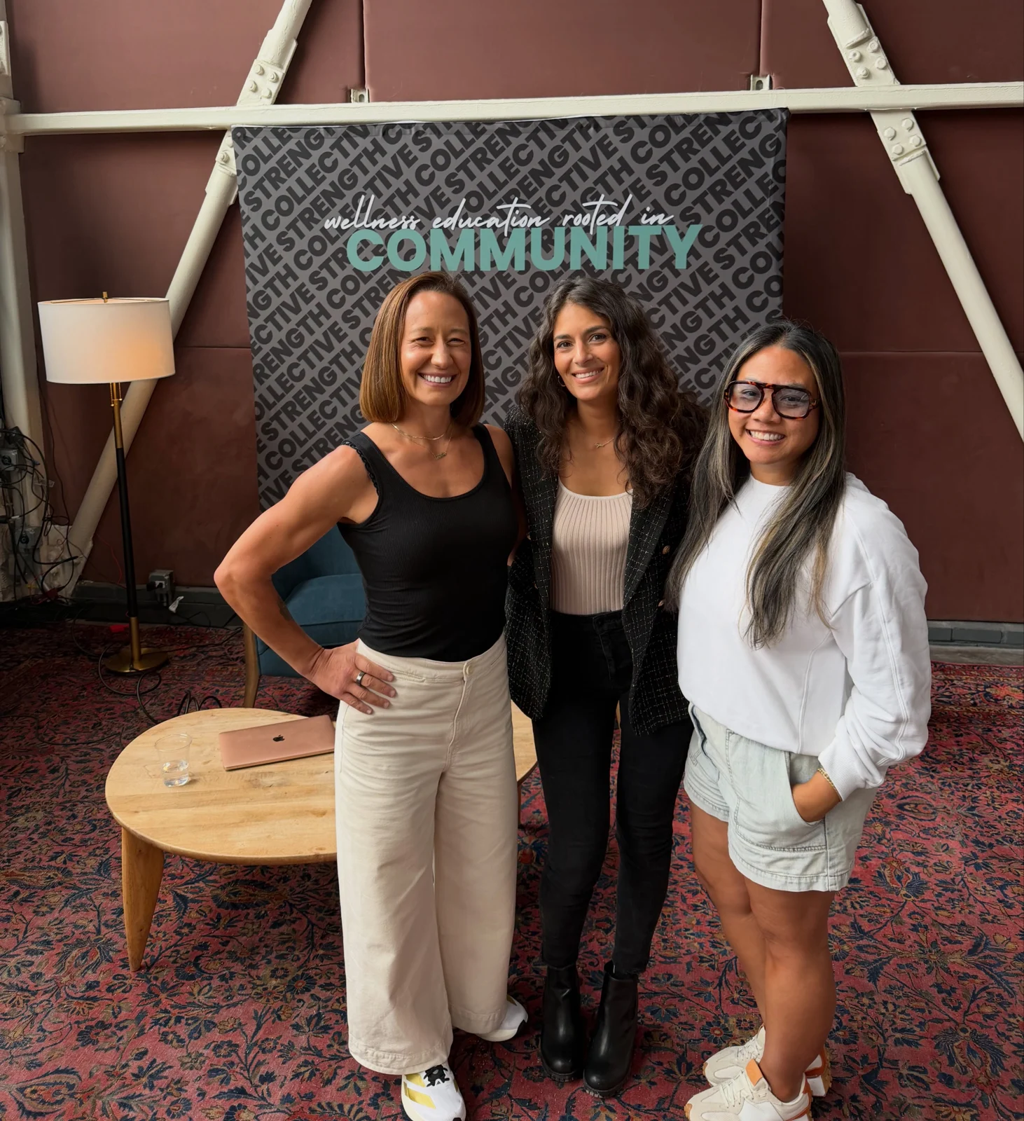 Three women smiling together in front of the Collective community backdrop.