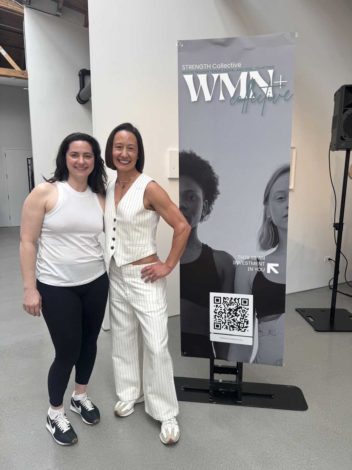 Two women smiling beside a WMN+ Collective banner.