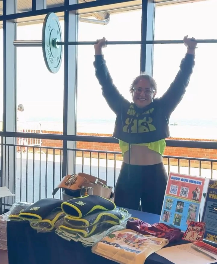 A smiling woman holding a barbell overhead behind a merchandise table.
