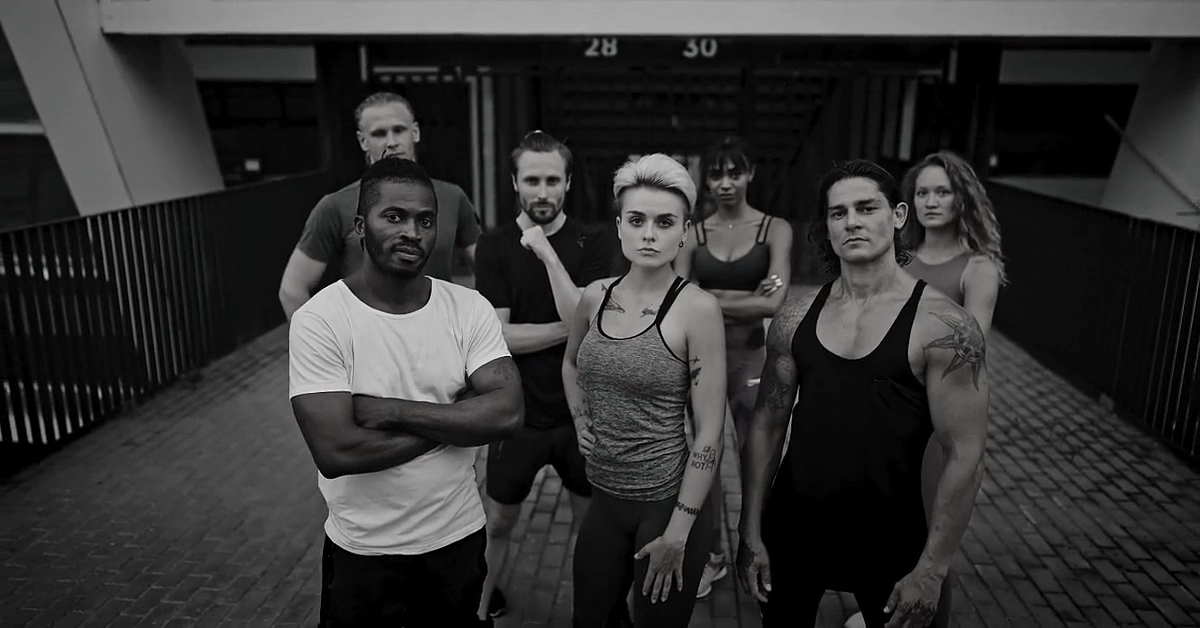 Seven fitness instructors in workout clothes standing together on a stadium walkway, in black and white.