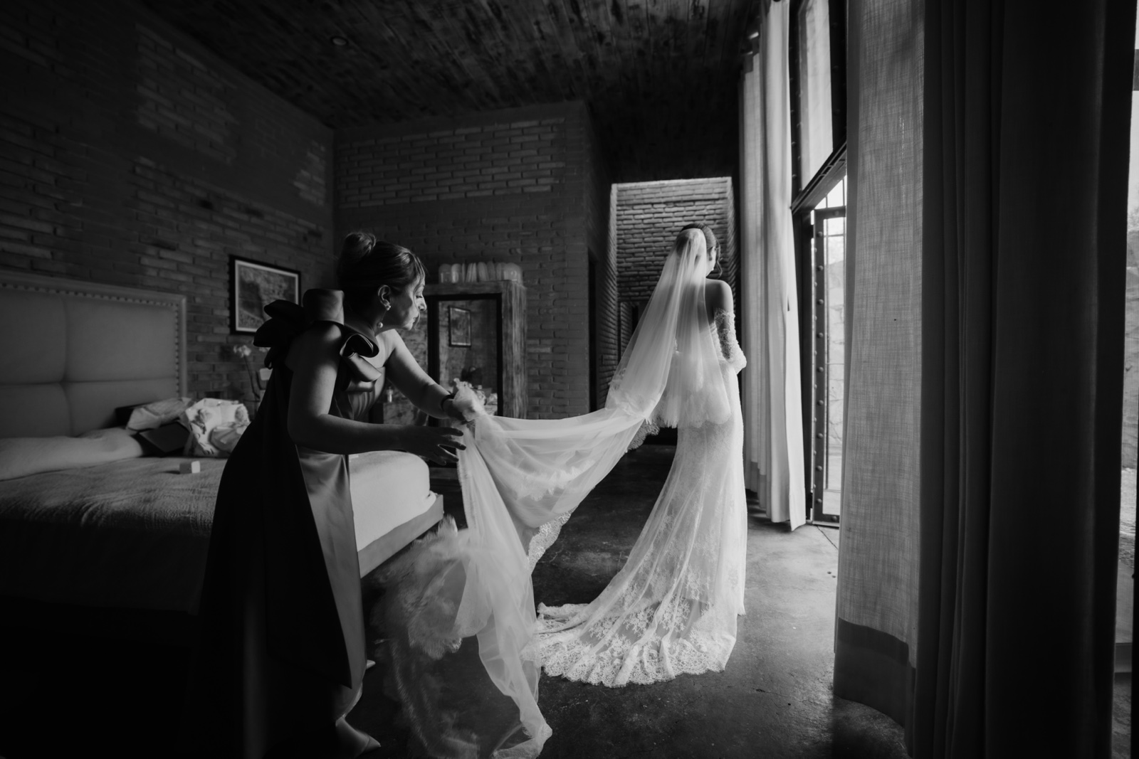 novia sosteniendo su vestido junto a una ventana, en blanco y negro