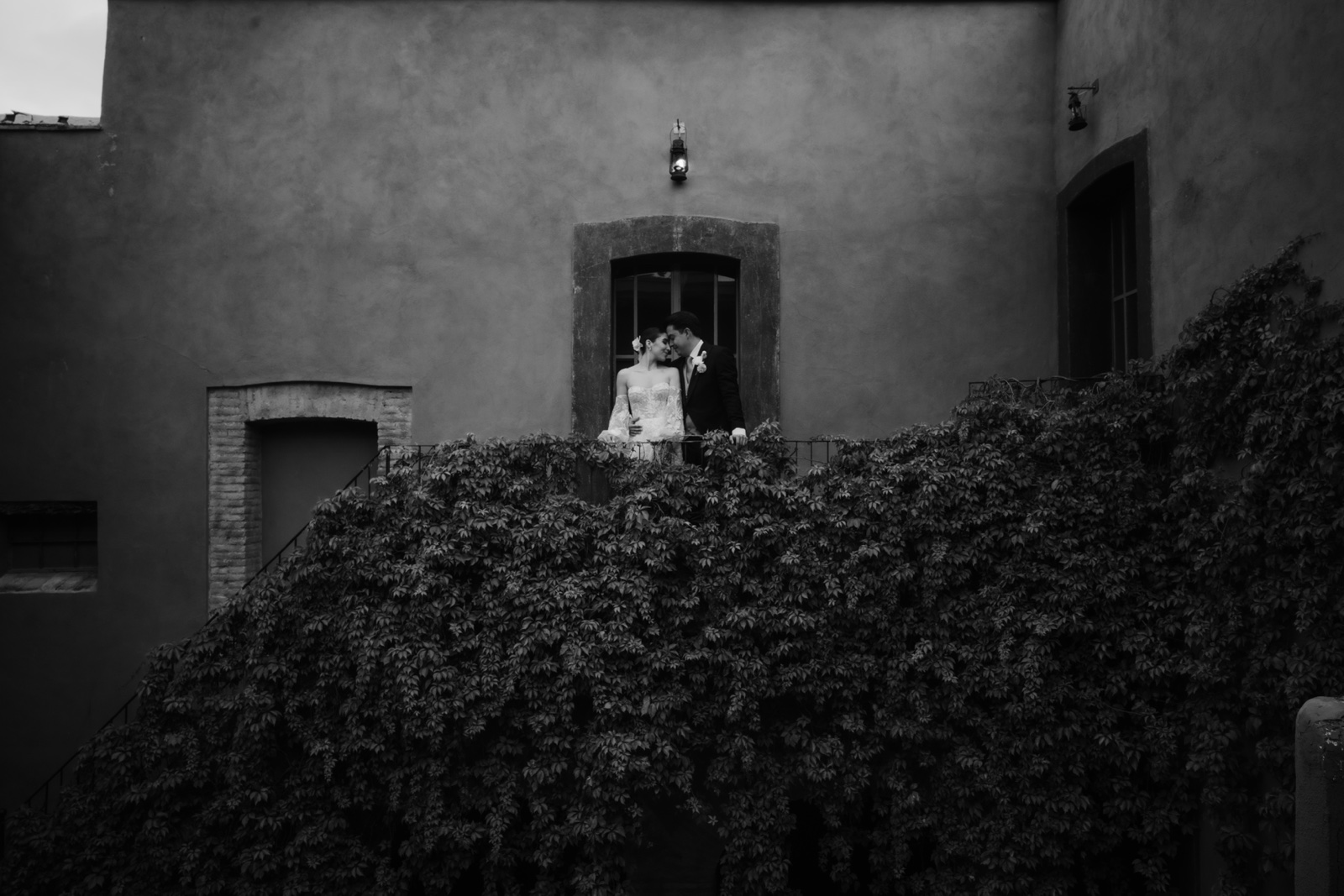 novios junto a una ventana sobre un muro cubierto de plantas, en blanco y negro