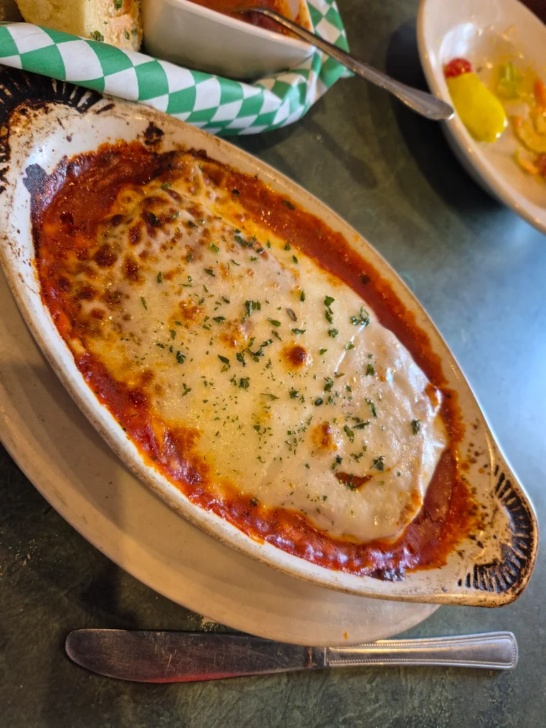 Baked Italian dish featuring melted cheese topped with herbs in a white, oval ceramic dish, served on a round beige plate with a knife below it. In the background, there is a basket with bread and a small bowl of sauce lined with green and white checkered paper, and a white bowl partially visible containing pickled vegetables.