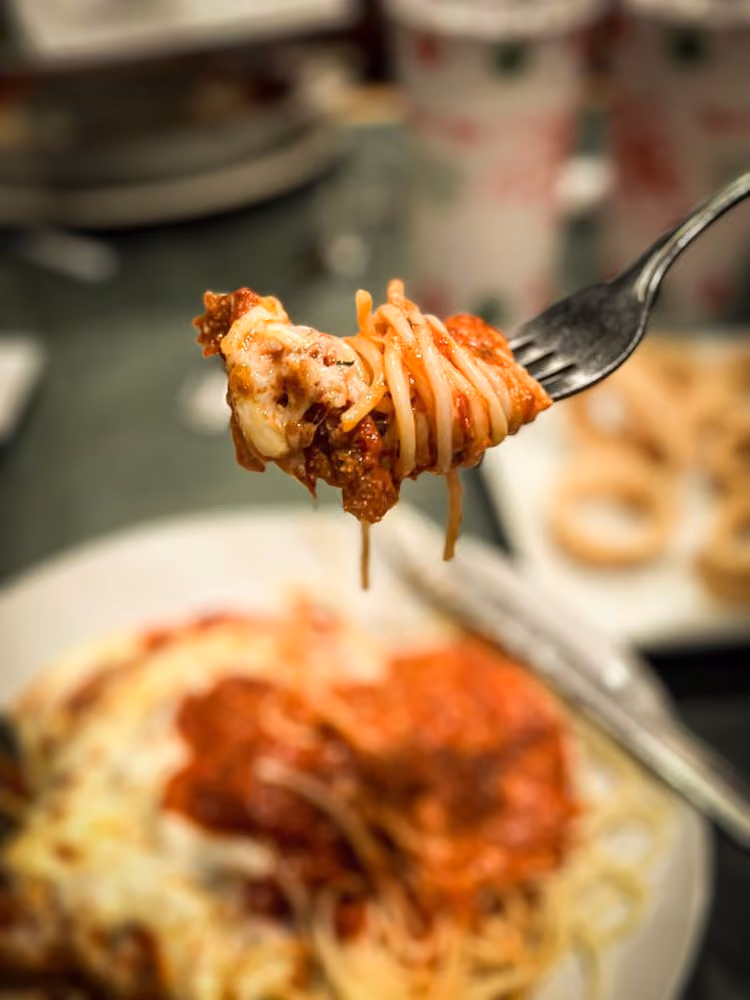 Close-up of a fork holding spaghetti pasta with meat sauce and melted cheese, with a plate of spaghetti in the background.