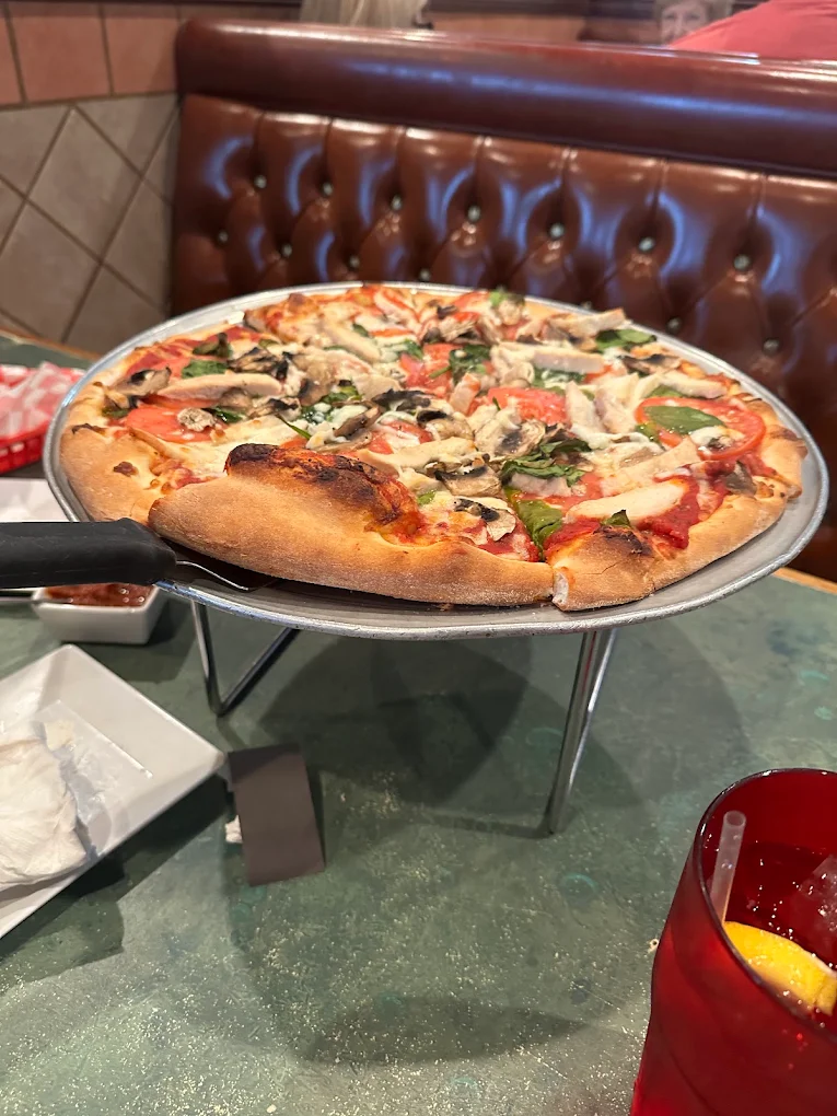 Fresh pizza on a metal stand with toppings including mushrooms, tomato slices, spinach, and chicken, placed on a restaurant table with a red drink and condiments nearby.