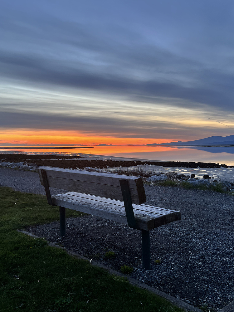 Empty bench on a gravel shoreline facing the water at sunset