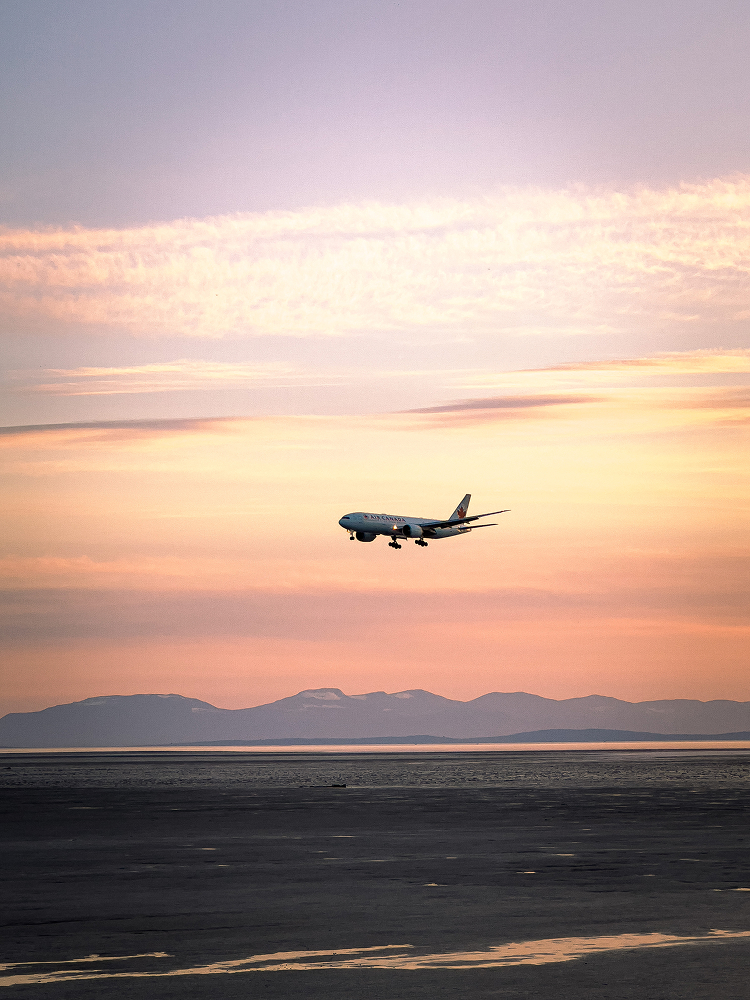 Airplane flying across a pink and orange sunset sky above distant mountains