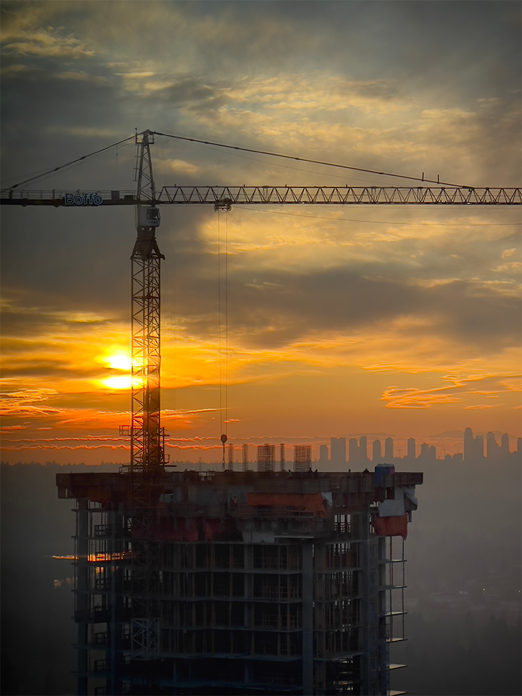 Construction crane rising over an unfinished tower at sunset with a hazy city skyline behind