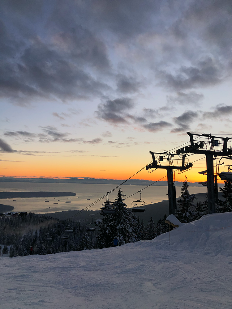 Ski chairlift towers above a snowy slope at sunset with the ocean and city far below