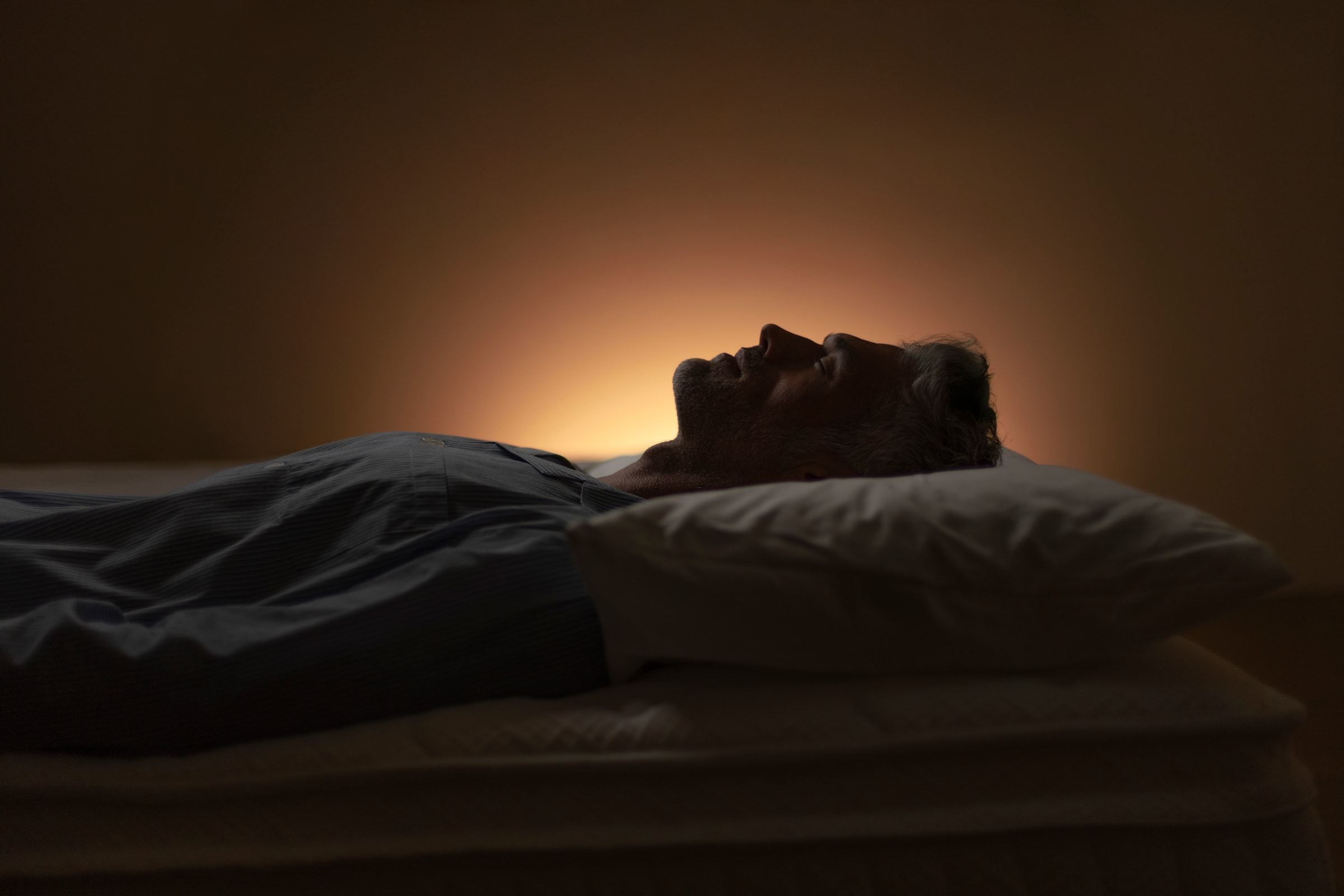 A sleeper lying on their back in low lamplight, head resting on a pillow