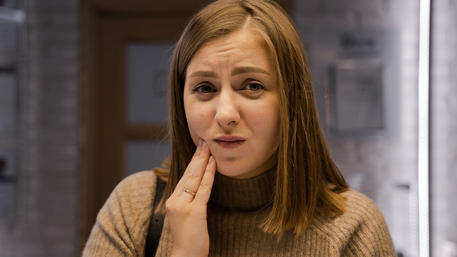 Caucasian woman expressing toothache discomfort, wearing a brown sweater indoors
