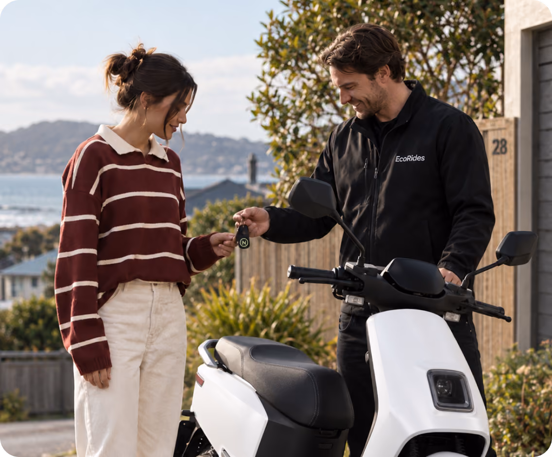 A man wearing a black jacket with 'EcoRides' hands over a key fob to a woman in a striped sweater next to a white electric scooter outdoors with houses and hills in the background.