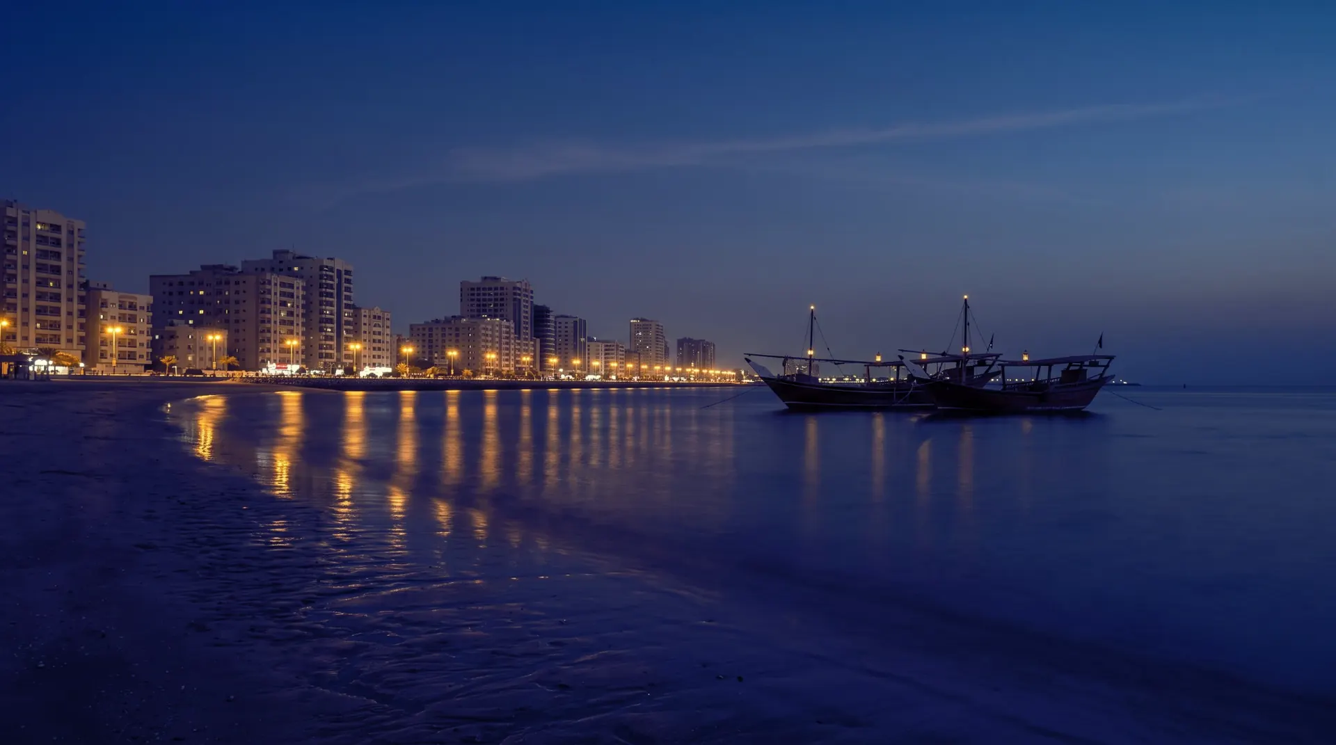 The Ajman corniche at blue hour, two wooden dhows moored on a calm sea below lit waterfront towers