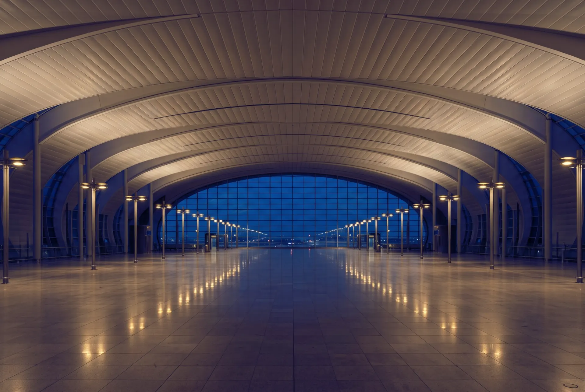 An empty airport departures hall at night under a long curved roof, the airfield beyond