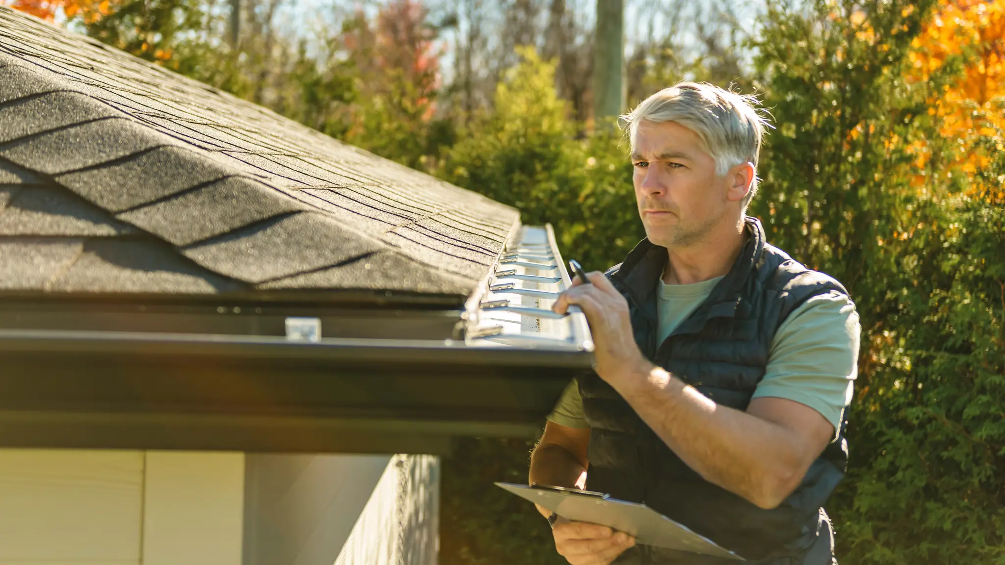 An adjuster inspecting a roof gutter and taking notes on a clipboard