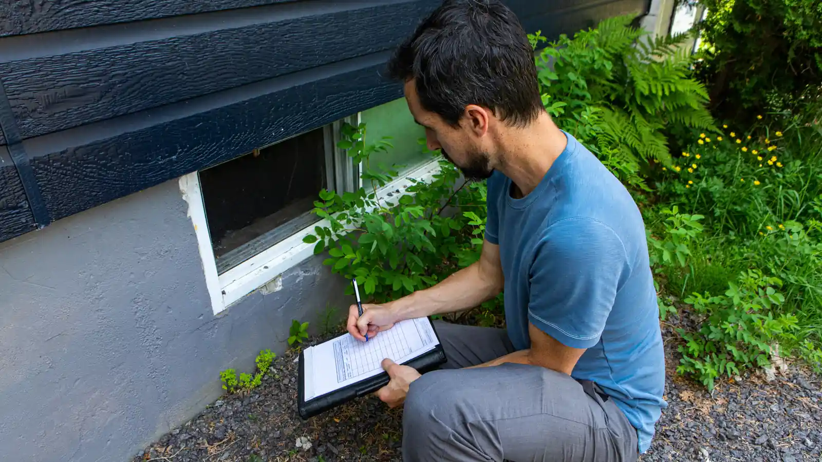 A public adjuster crouched by a home's foundation window, writing inspection notes on a clipboard